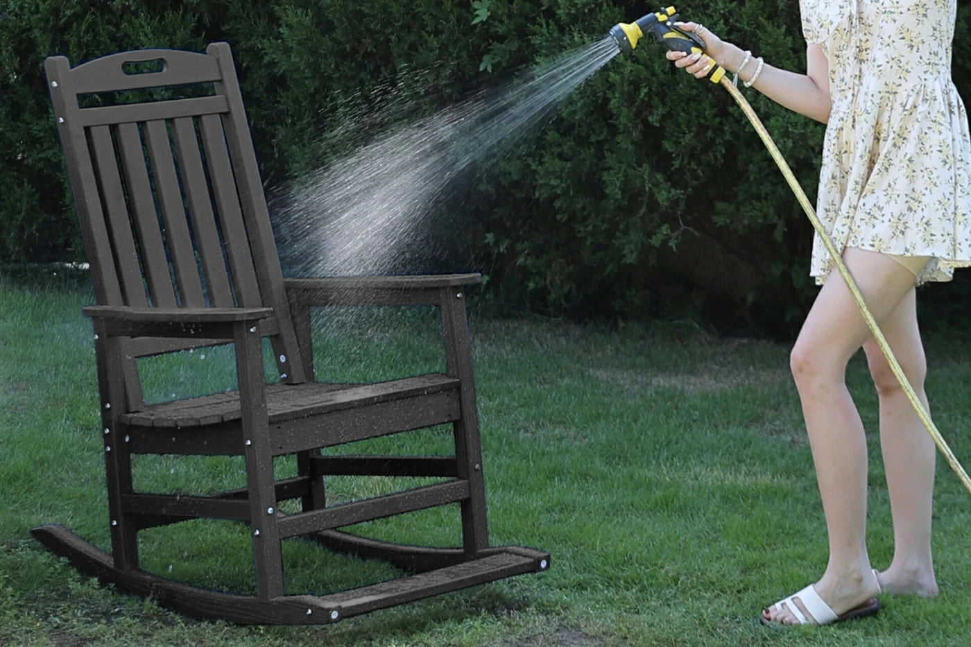 Person cleaning a wooden rocking chair with a hose on a grassy area.