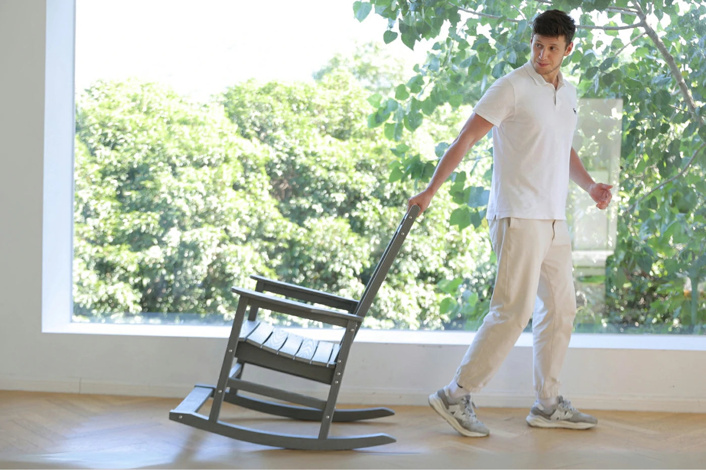 Person moving a rocking chair indoors with large windows showing greenery outside.