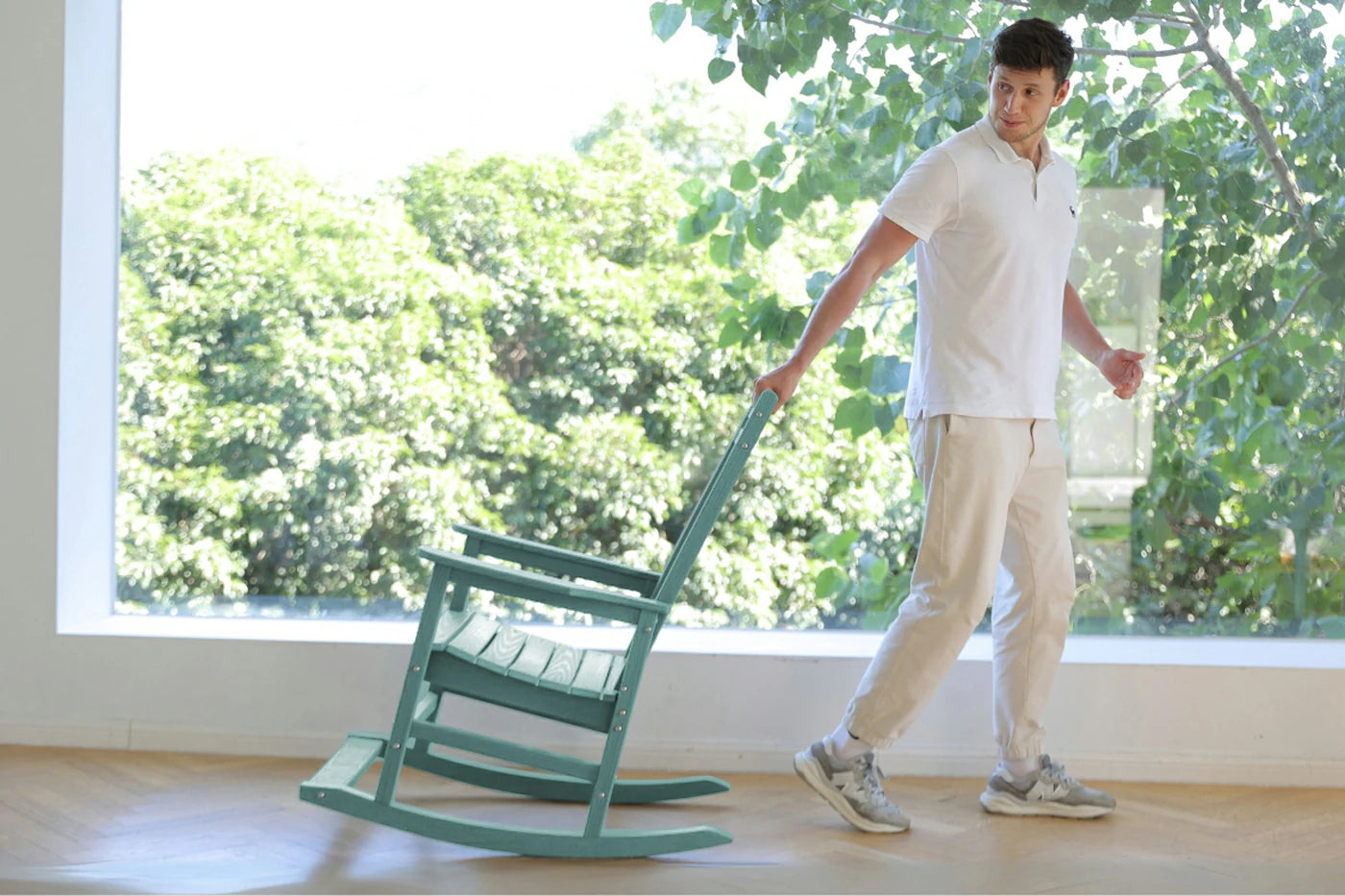 Person pushing a green rocking chair indoors with a large window showing greenery outside.