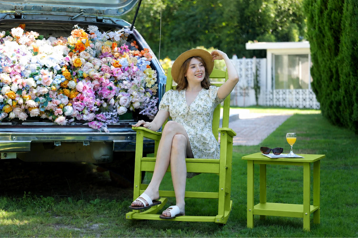 A woman in a floral dress and straw hat relaxes in an apple green wooden rocking chair beside an open car trunk filled with colorful artificial flowers, with a matching green side table holding sunglasses and an orange drink on a grassy lawn.