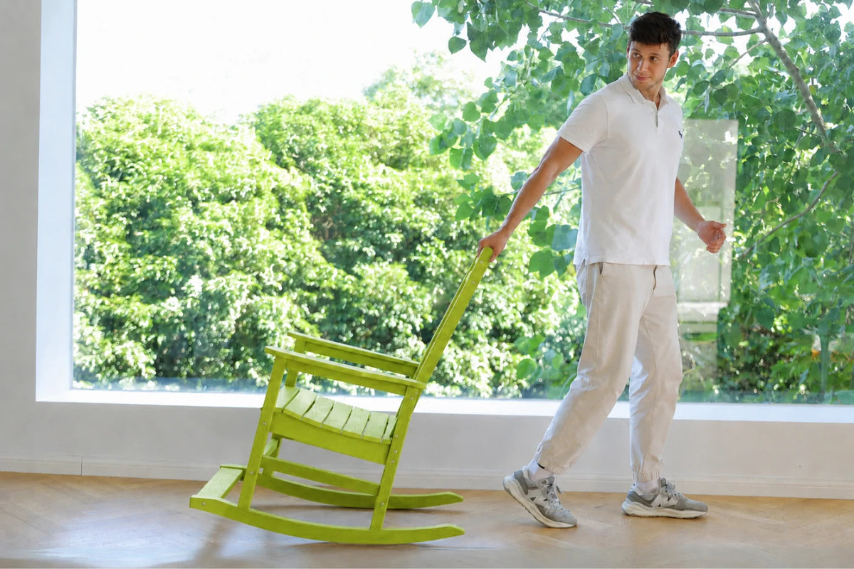 A man in a white polo shirt and light pants pushes an apple green wooden rocking chair with a pull handle inside a room with a large window showing green trees outside.