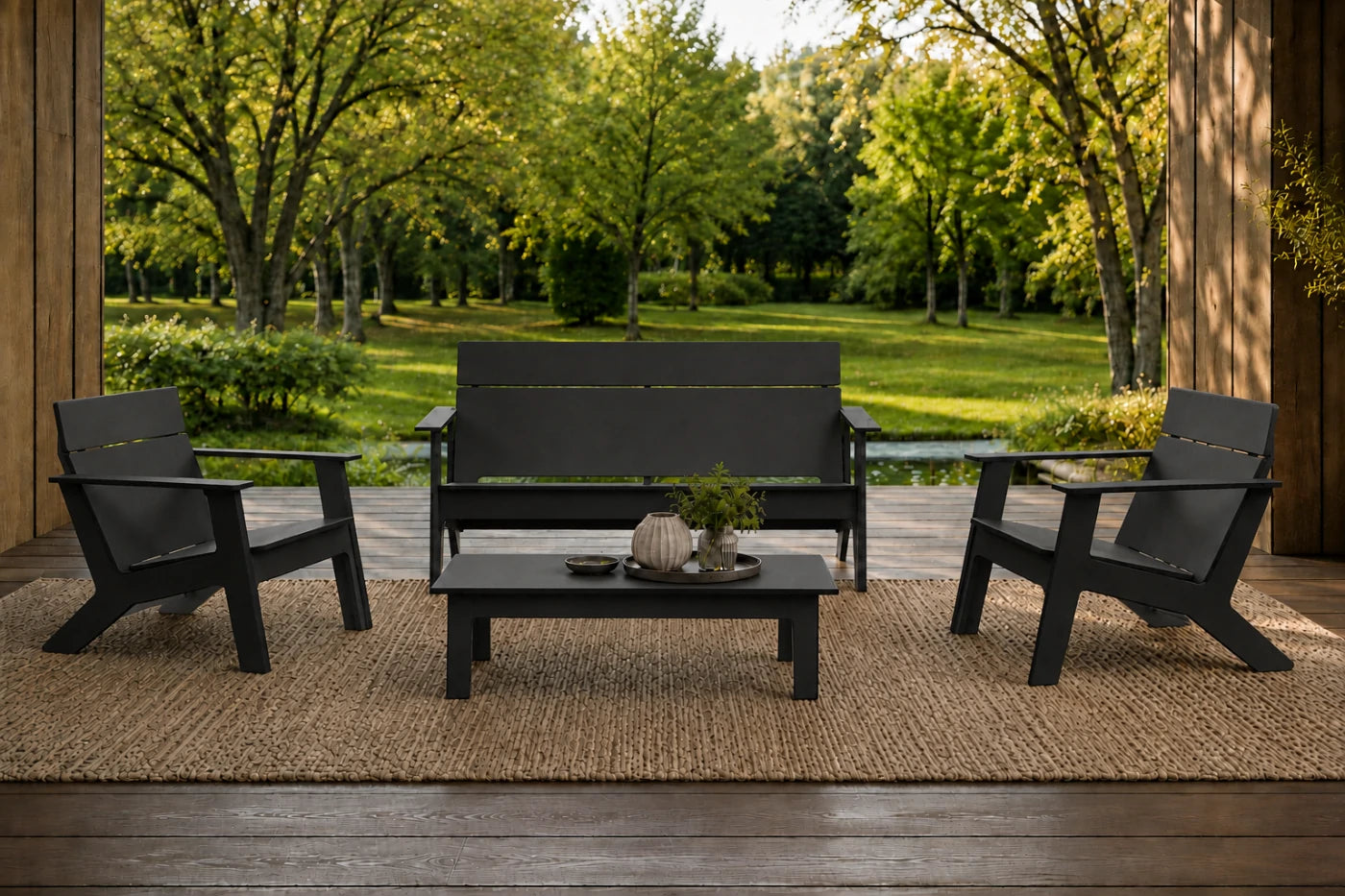A 4-piece outdoor patio furniture set including a loveseat, two armchairs, and a coffee table, all in a black finish, arranged on a woven jute rug on a wooden porch with a lush green garden background.