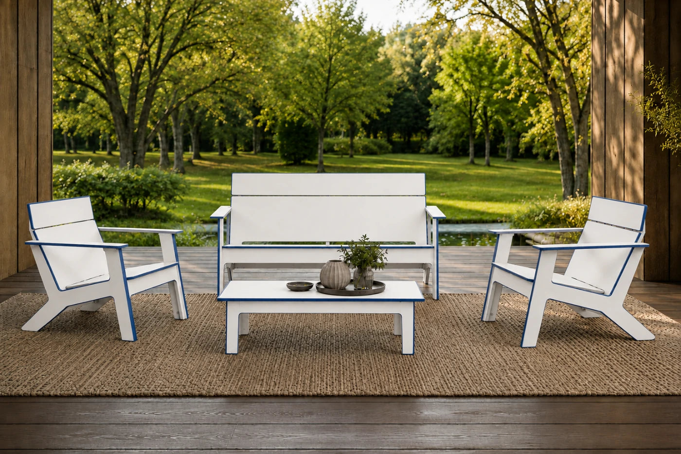 A 4-piece outdoor patio furniture set including a loveseat, two armchairs, and a coffee table, all in white with blue trim, arranged on a jute rug on a wooden deck with a lush green garden background.