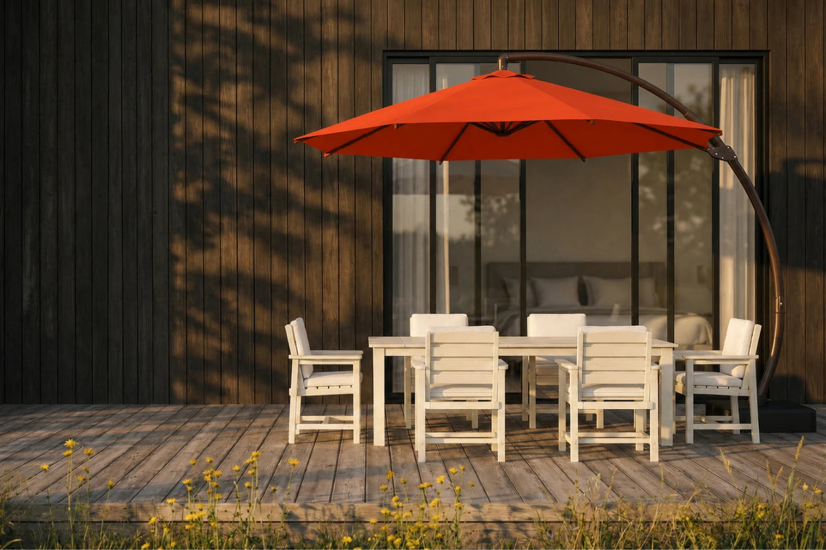 An orange patio umbrella with a curved dark metal arm stands over a white outdoor dining table and chairs on a wooden deck in front of a modern house with dark wood siding.