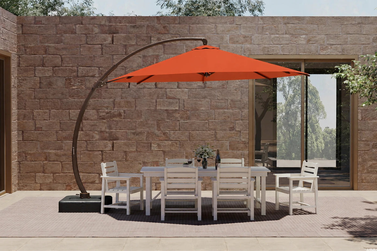 A large outdoor patio setup featuring an orange cantilever umbrella over a white dining table and six matching chairs, set against a stone wall background.