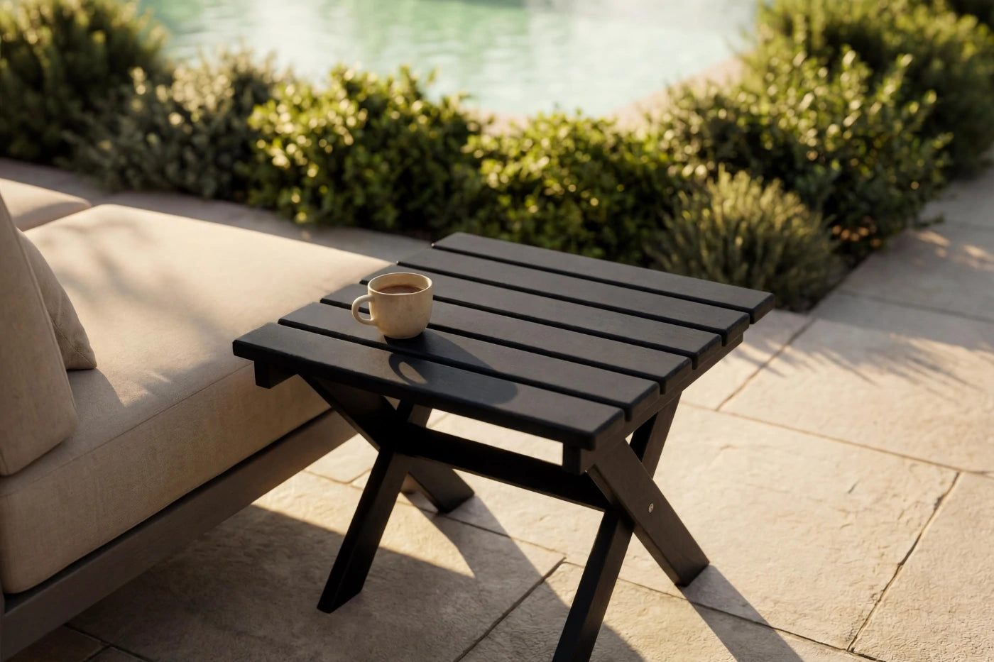 Black outdoor side table with a cup on it, next to a beige sofa by a pool.