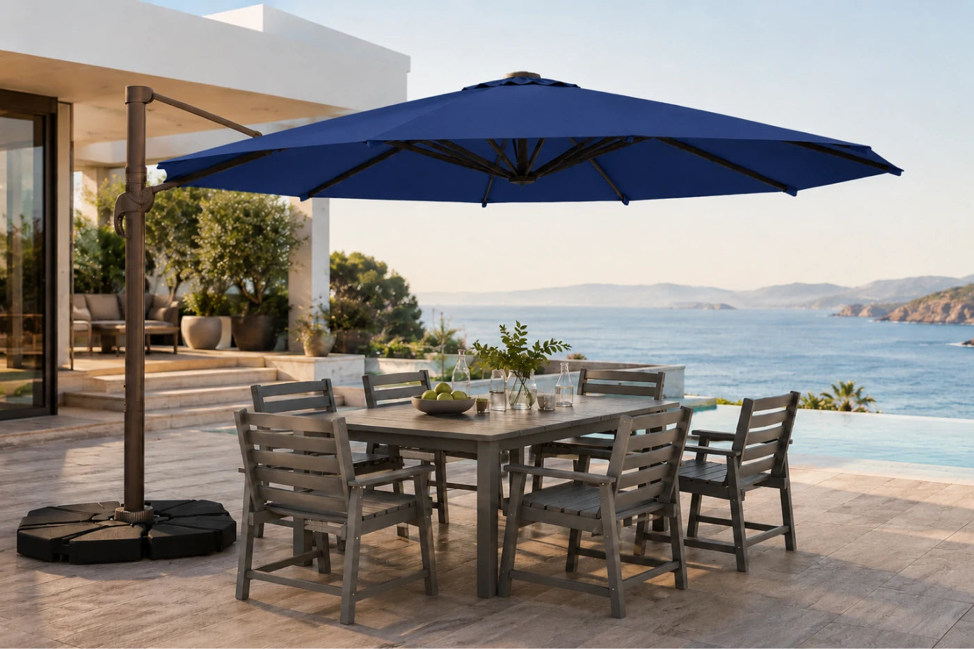 A large patio umbrella with a black cantilever pole and weight base, shading a gray outdoor dining table and six chairs on a stone terrace overlooking a blue ocean and sea walls.