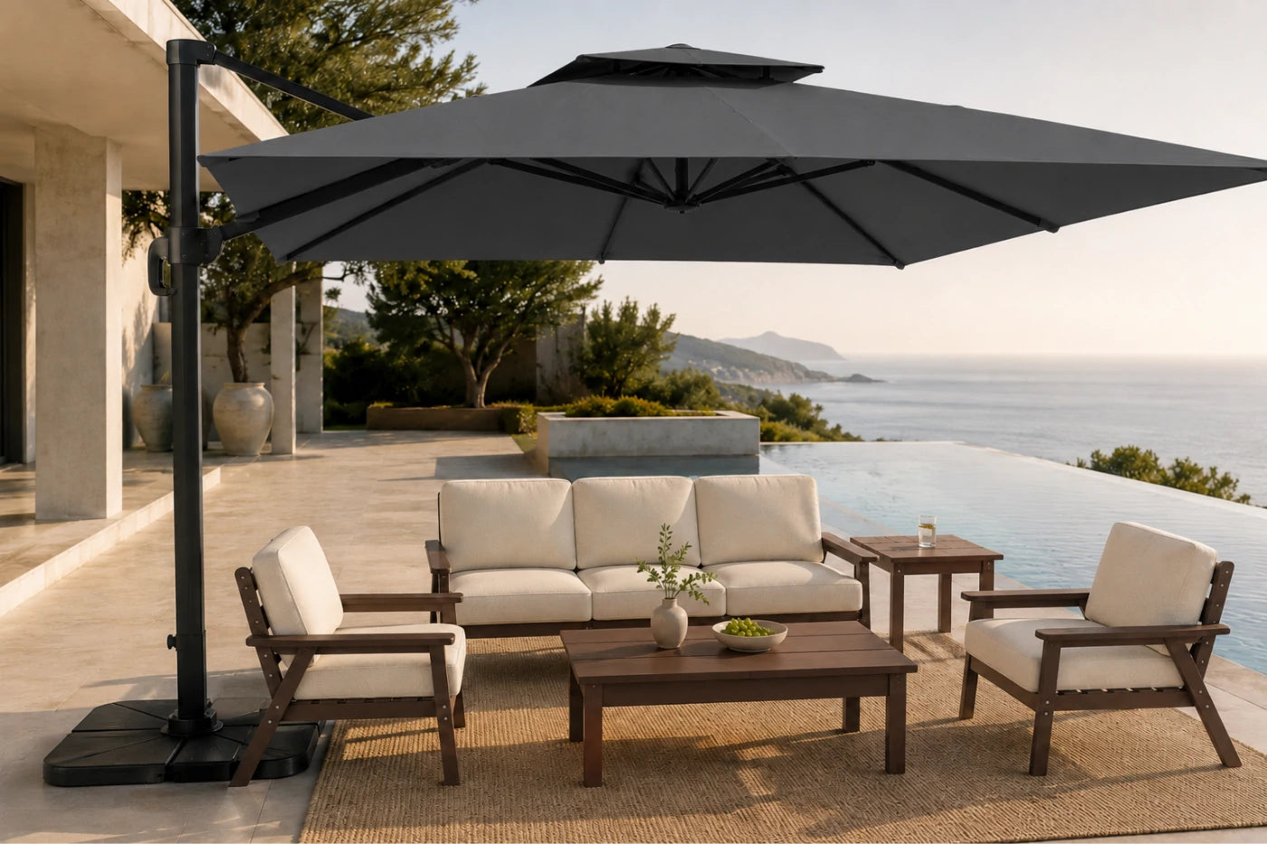 A large dark gray patio umbrella with a cantilevered pole stands over an outdoor seating area on a sunlit terrace overlooking a pool and sea. The furniture includes a cream cushioned sofa, two matching armchairs, a wooden coffee table, and a side table.