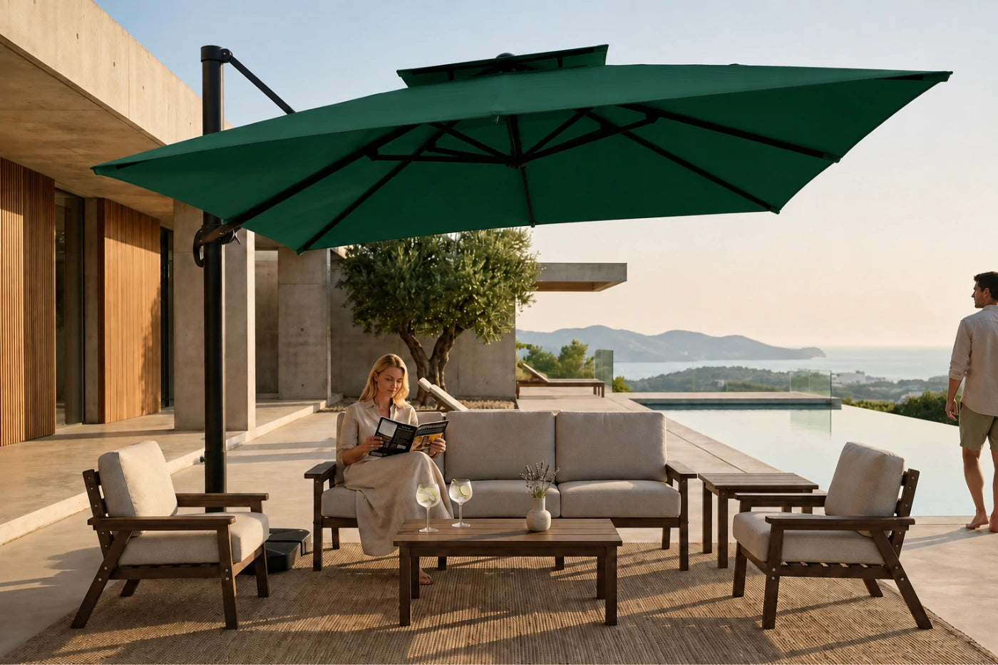 Outdoor patio furniture set with a large dark green cantilever umbrella, a three-seat sofa, two armchairs, and a coffee table on a poolside terrace. A woman sits on the sofa reading, and a man stands nearby, with a modern building and distant hills in the background.
