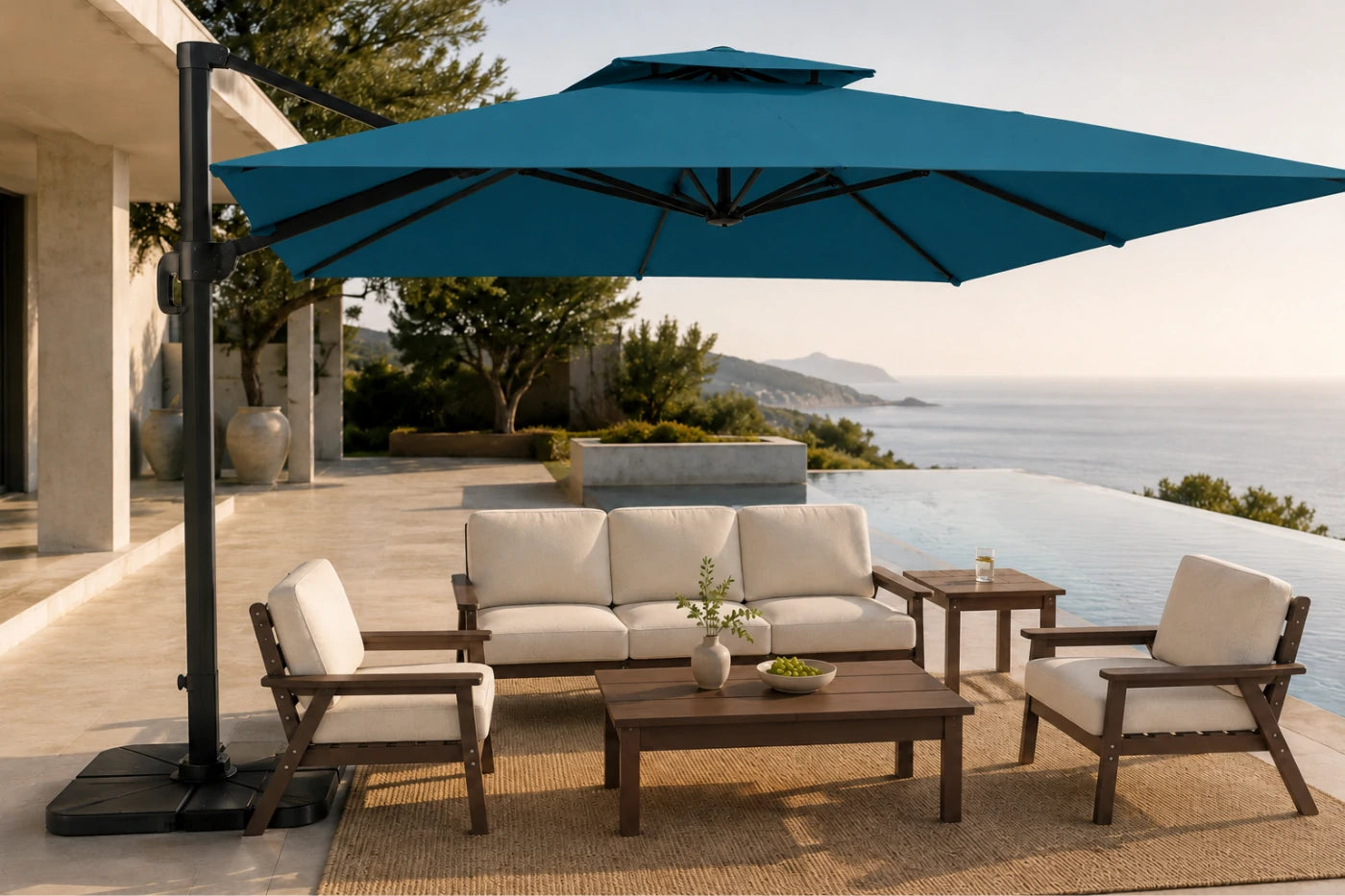 Outdoor patio furniture set with a large lake blue cantilever umbrella, a wooden three-seat sofa, two matching armchairs, and a coffee table on a woven rug beside an infinity pool and ocean view.