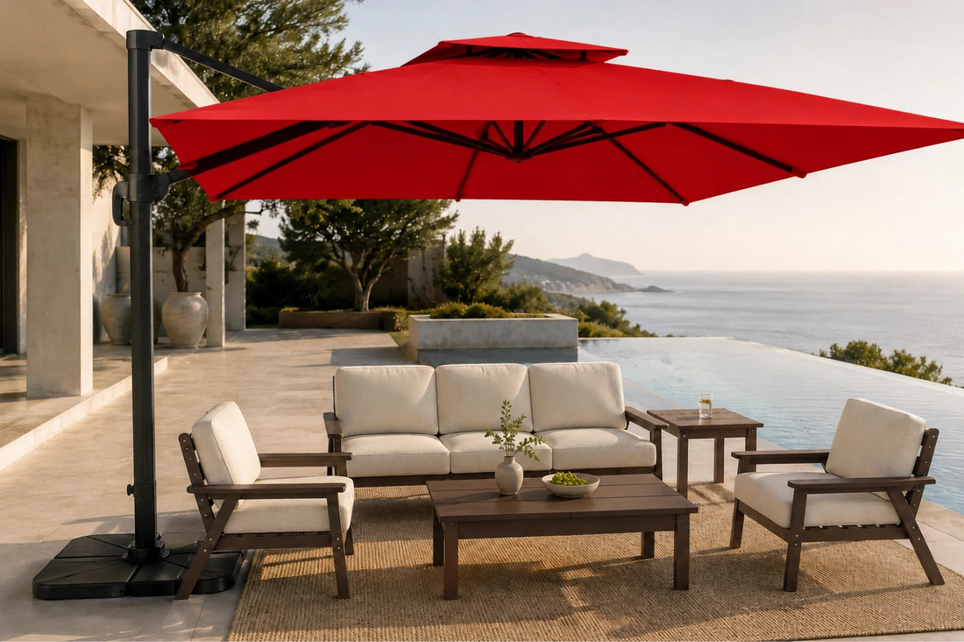 A luxury outdoor patio setup featuring a large red cantilever patio umbrella, a three-seater sofa, two armchairs, a coffee table, and a side table on a woven rug, set against an infinity pool and ocean backdrop.
