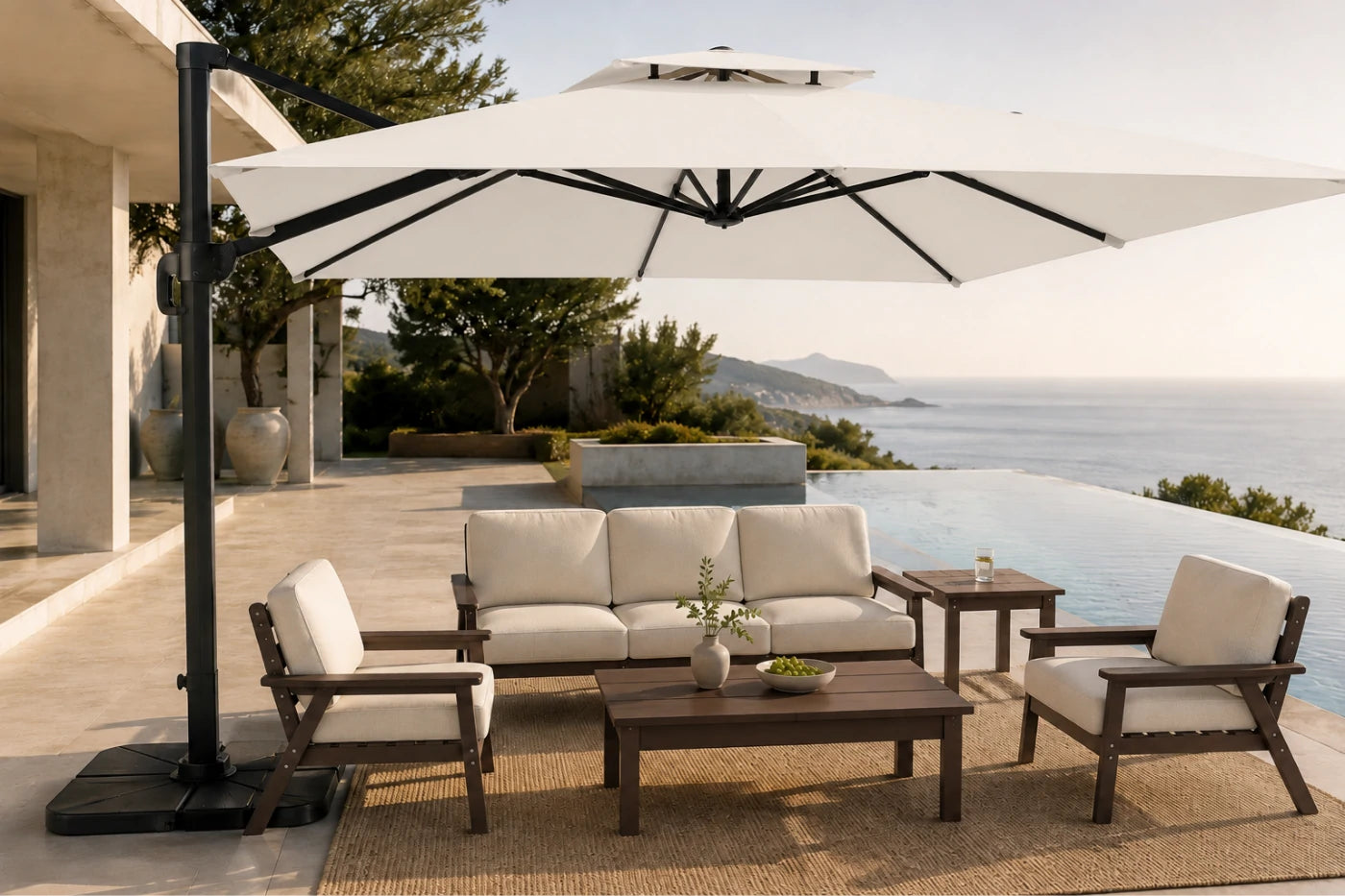 Outdoor patio furniture set with a large white cantilever umbrella, a three-seat sofa, two armchairs, a coffee table, and a side table on a tiled deck overlooking an infinity pool and sea.