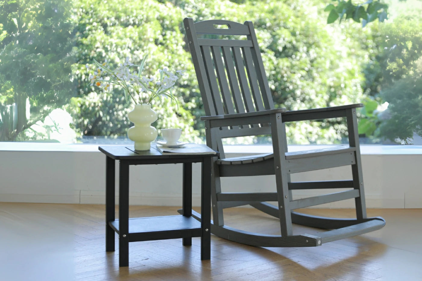 Gray rocking chair with a small table on a wooden floor with a blurred green outdoor background