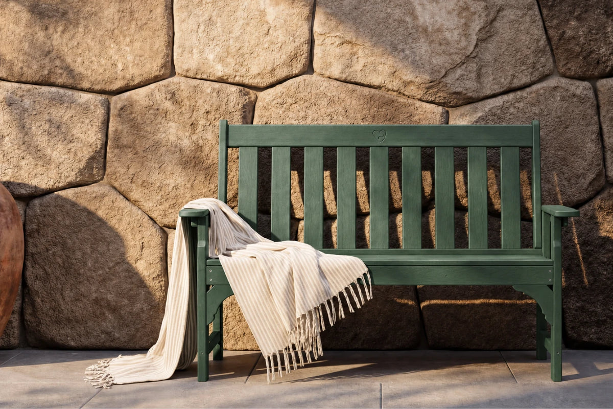 A dark green bench with a cream-colored fringed throw blanket draped over it, set against a rustic stone wall.