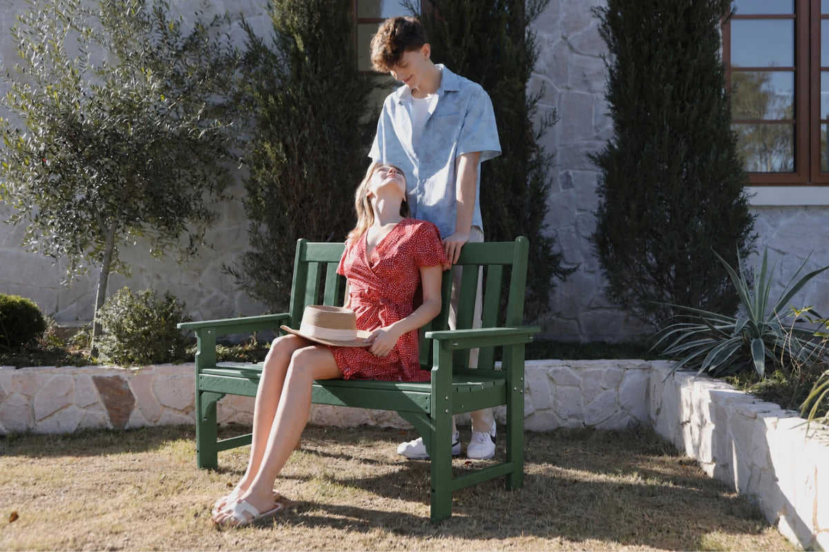 A man in a light blue shirt stands behind a woman wearing a red dress seated on a dark green bench in a garden setting.