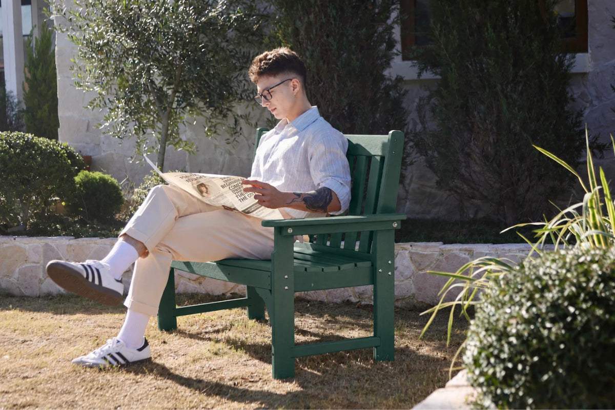 A man wearing glasses sits on a dark green outdoor wooden bench reading a newspaper, dressed in a white shirt, beige trousers, white socks, and white sneakers, set against a sunny garden background.