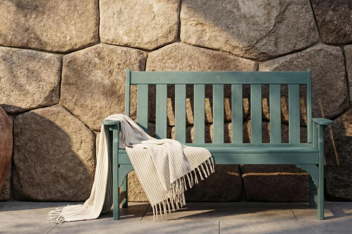 Green bench with a blanket draped over it against a stone wall.