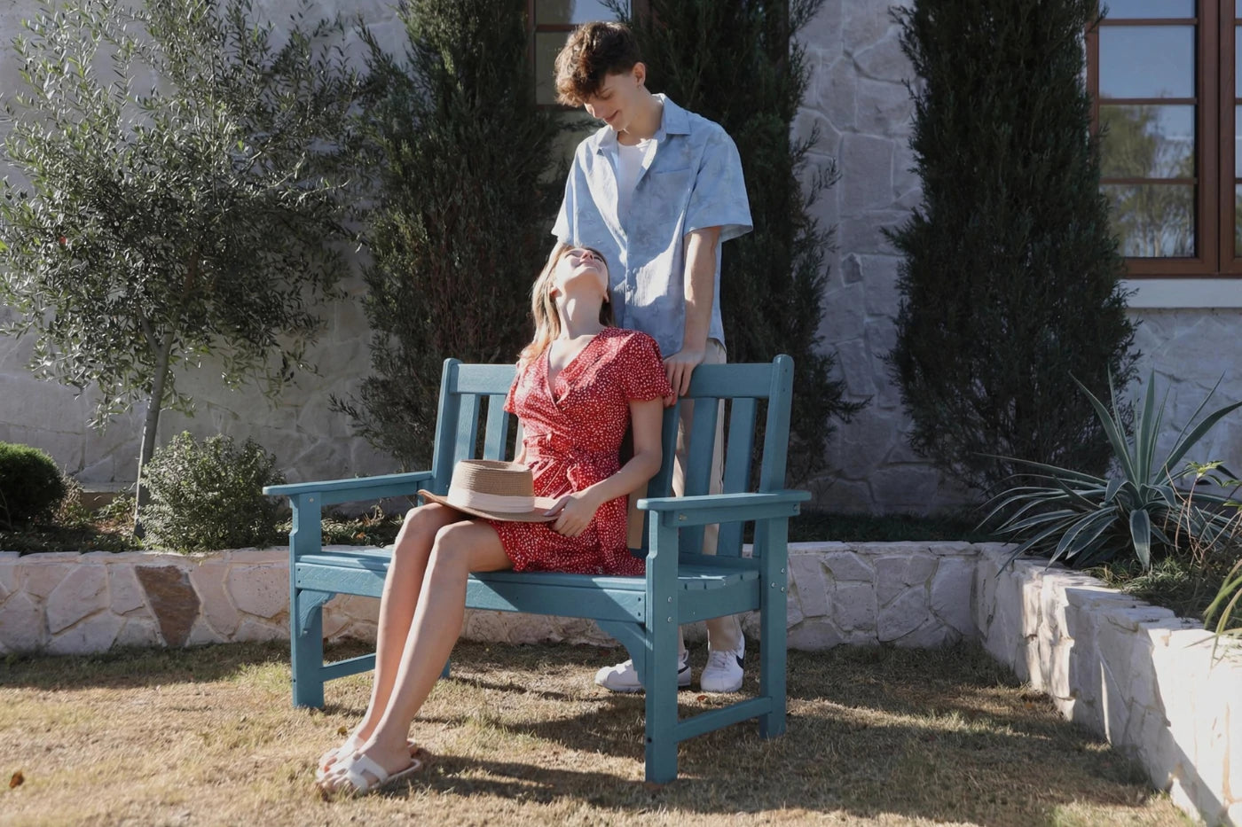 Two people sitting on a blue bench outdoors with a stone wall and trees in the background.