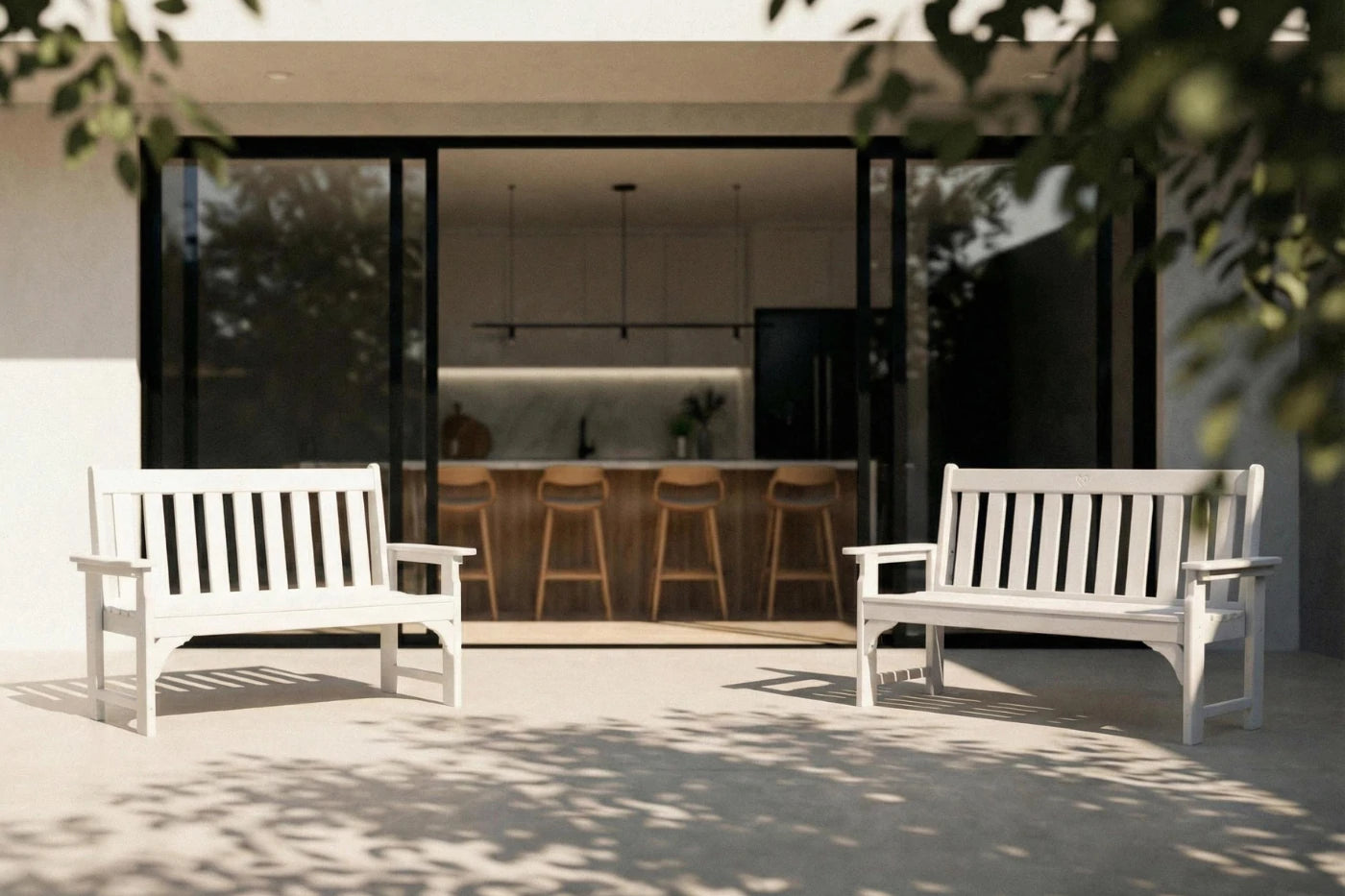 Two white benches on a patio in front of a modern house with a kitchen visible through large windows.