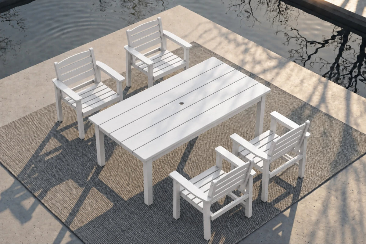 White outdoor dining set consisting of a rectangular slatted table and four matching armchairs, arranged on a textured gray rug beside a pool with water and tree reflections visible.