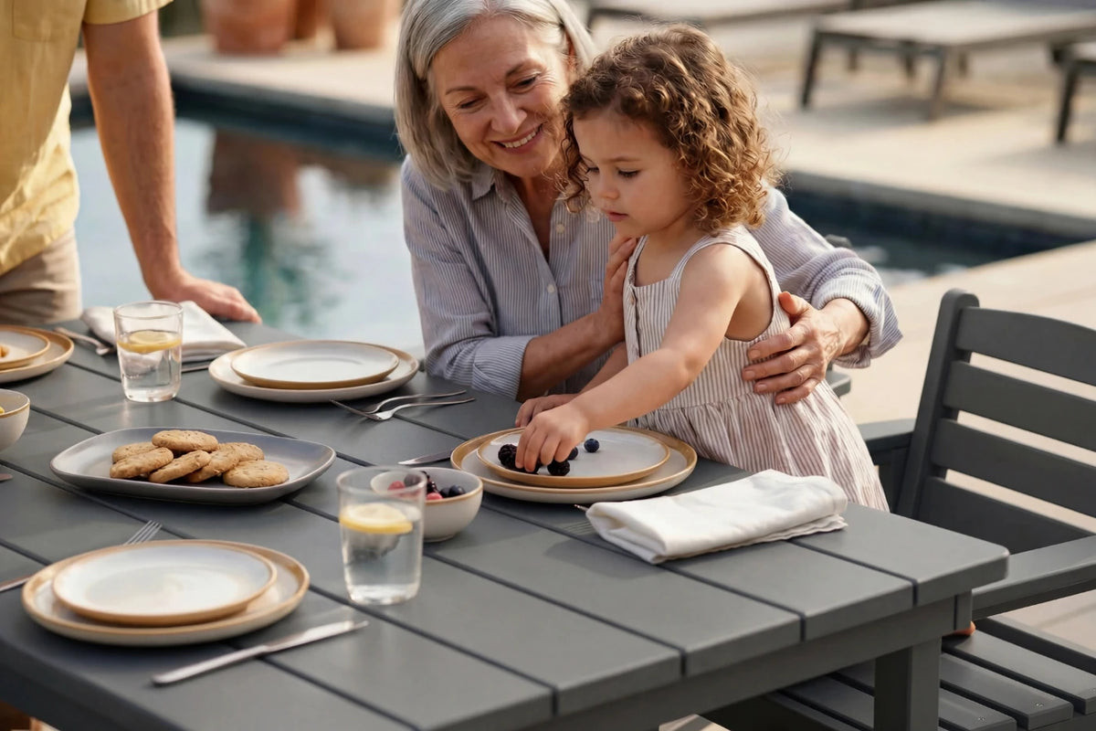 A grandmother and young daughter share a meal at an outdoor patio table by a swimming pool. The table is set with plates, glasses, and a folded napkin.