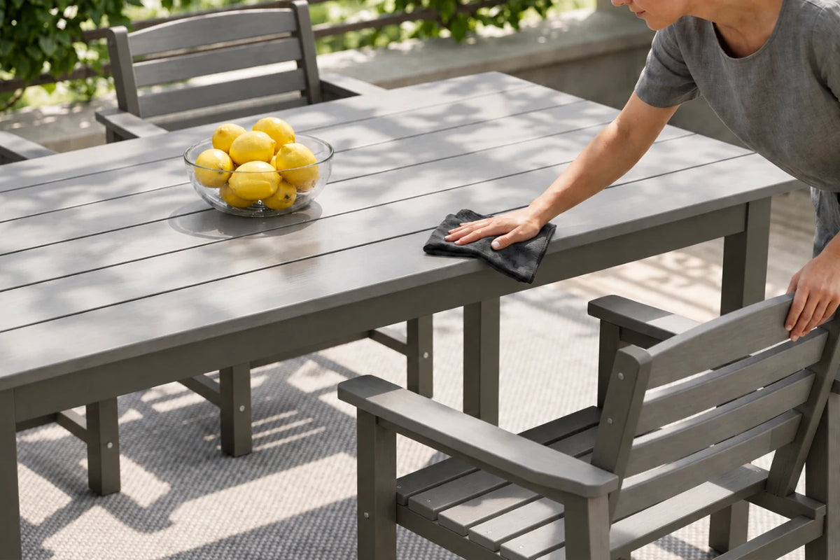 A person outdoors wipes down a gray slatted outdoor dining table with a dark cloth. A glass bowl of lemons sits on the table, with matching gray chairs around it.