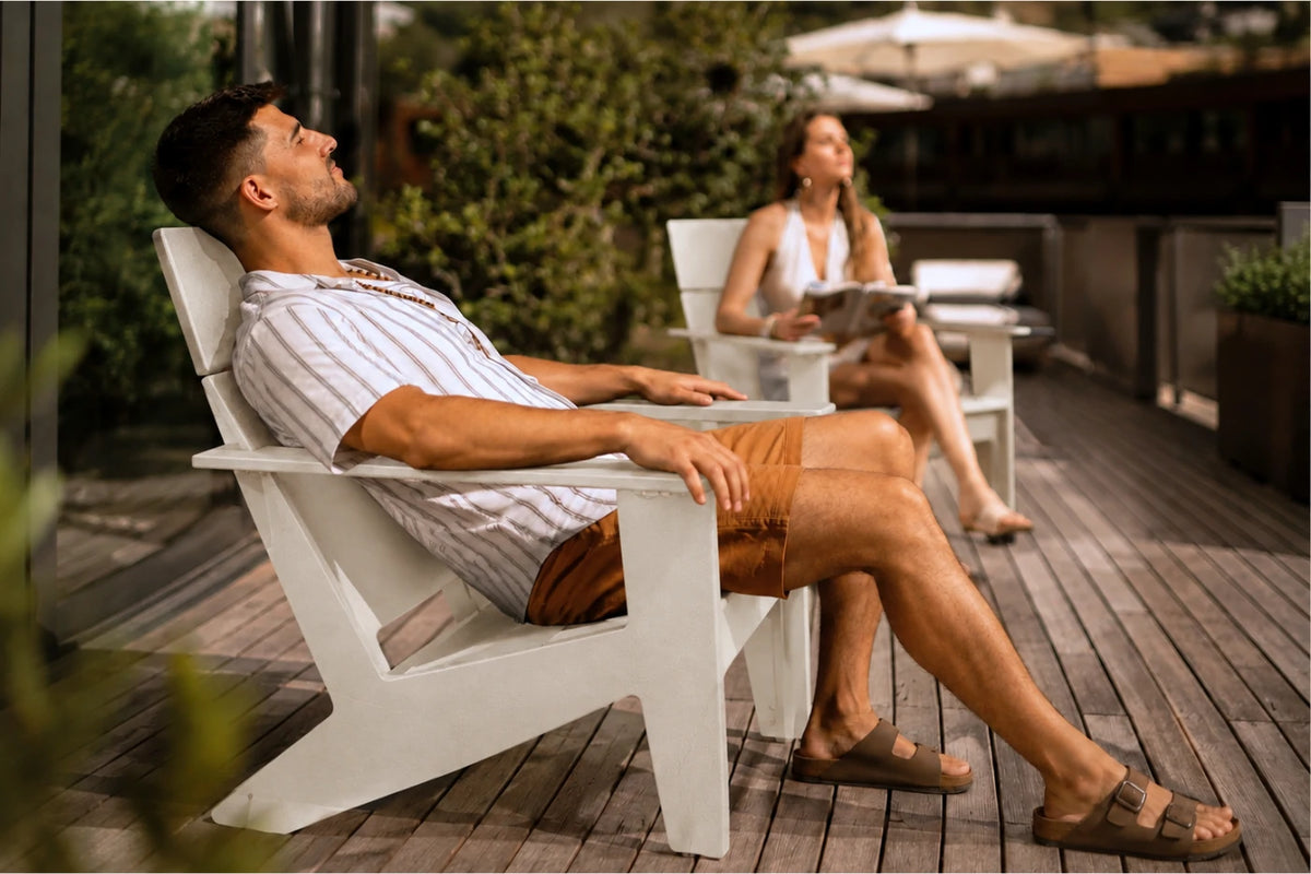A man relaxes in a white outdoor Adirondack chair on a wooden patio, wearing a striped shirt, brown shorts, and sandals. A woman sits in a matching chair in the background.