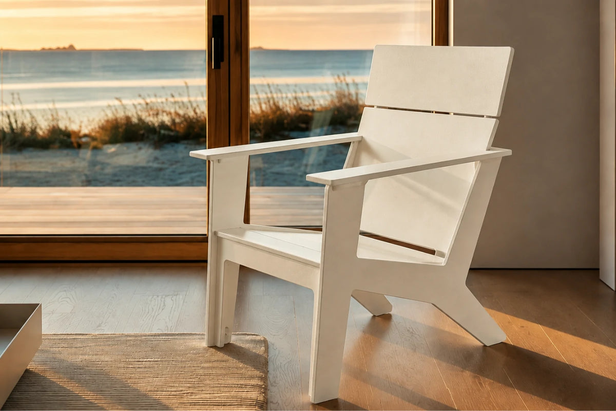 A modern white outdoor Adirondack chair positioned on a wooden deck in front of large glass doors overlooking a beach at sunset.