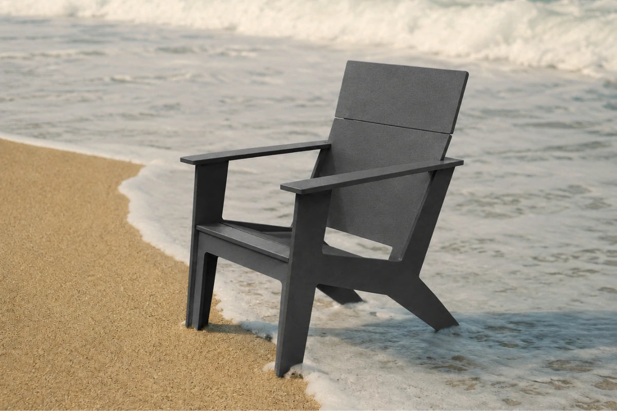 A dark gray modern outdoor chair positioned on a sandy beach with ocean waves washing onto the shore in the background.