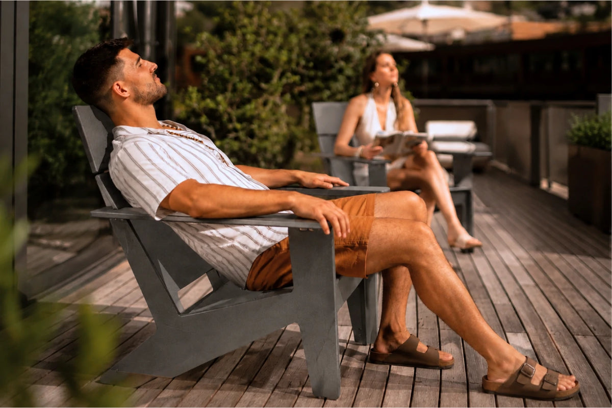 A man relaxes in a dark gray Adirondack chair on a wooden deck, wearing a white striped t-shirt, brown shorts, and brown sandals. A woman sits in a blurred chair in the background on the patio.