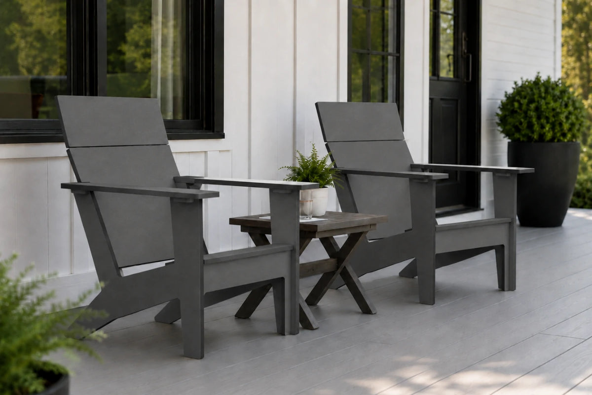 Two dark gray outdoor Adirondack chairs with a small wooden side table between them, set on a light porch in front of a white house with black-framed windows.
