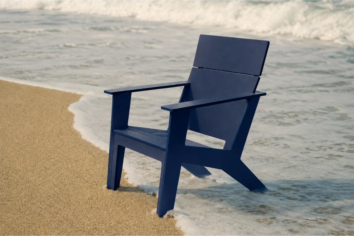 A navy blue Adirondack-style chair placed on a sandy shore with ocean waves washing around it.