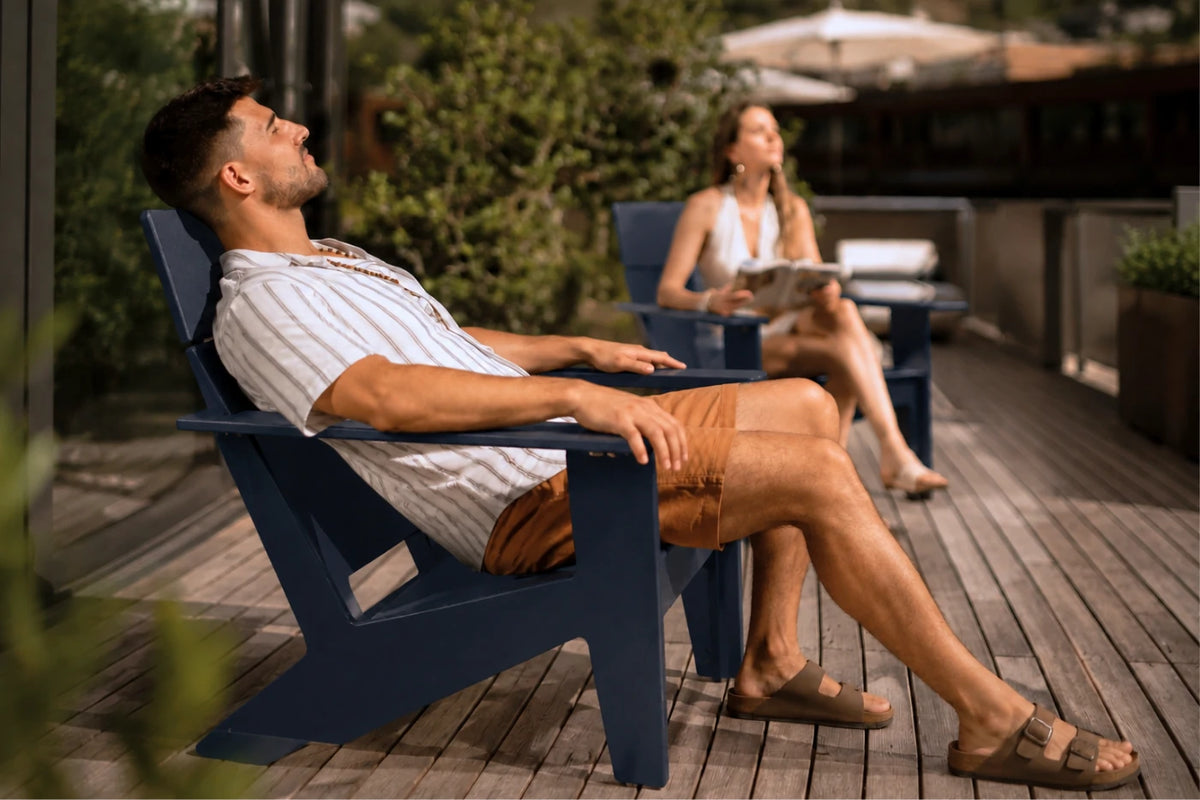 A man relaxes in a navy blue outdoor Adirondack chair on a wooden deck, wearing a white striped shirt and brown shorts. A woman sits in a matching chair in the background.