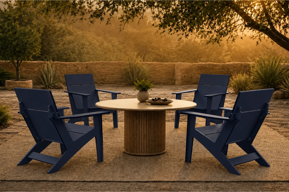 A set of four navy blue outdoor Adirondack-style chairs arranged around a round light wood dining table on a patio, with trees and warm golden evening light in the background.