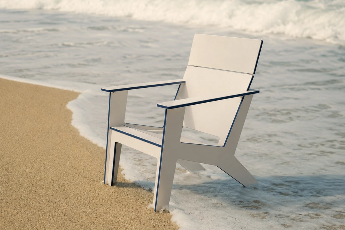 A modern white outdoor chair with dark blue trim sits on a sandy beach with ocean waves in the background.