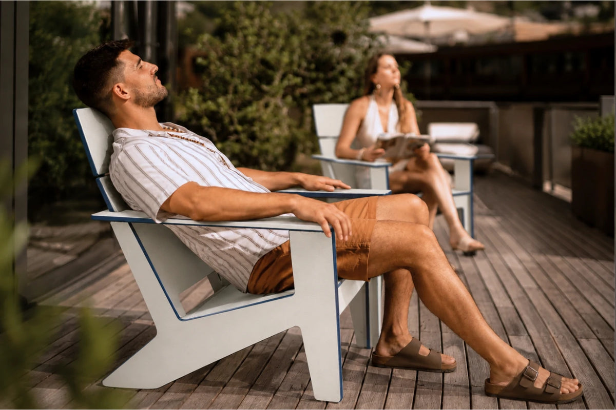 A man relaxes in a modern white outdoor chair with blue trim on a wooden deck, with a woman seated in a blurred chair in the background.