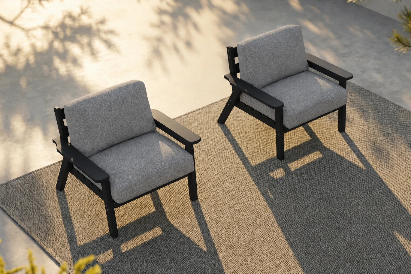 Two outdoor armchairs with gray textured cushions and black frames, positioned on a textured gray patio rug with warm sunlight and leaf shadows.
