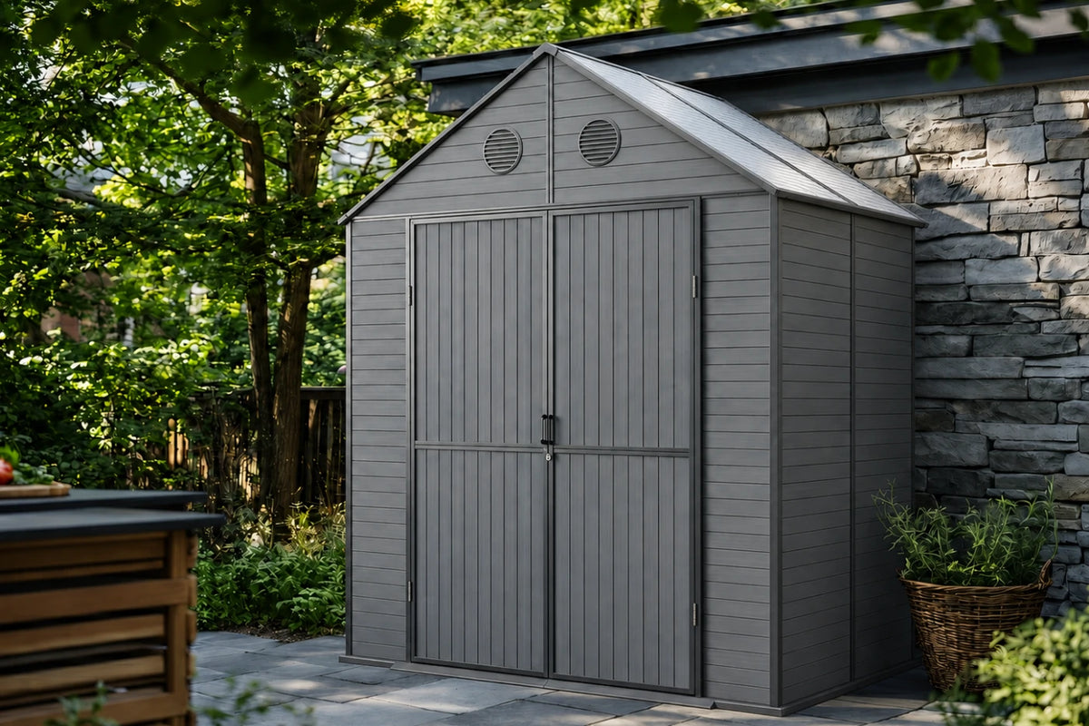 A gray resin storage shed with polycarbonate roof, shown outdoors next to a stone wall and green trees.
