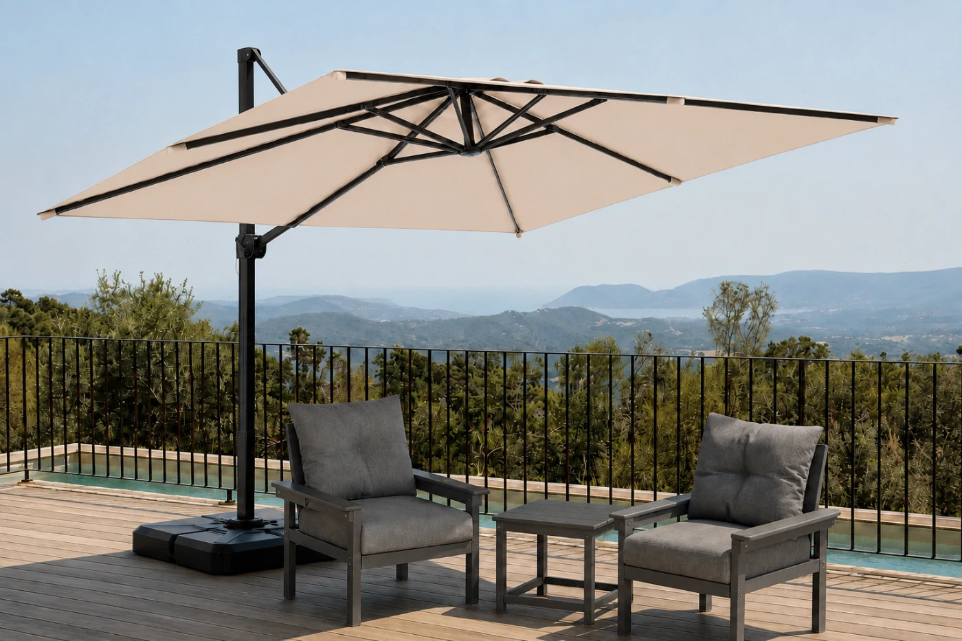 A large beige patio umbrella with a black cantilever pole and base, shown on a wooden deck with two gray outdoor armchairs and a small side table, overlooking a scenic mountain landscape.
