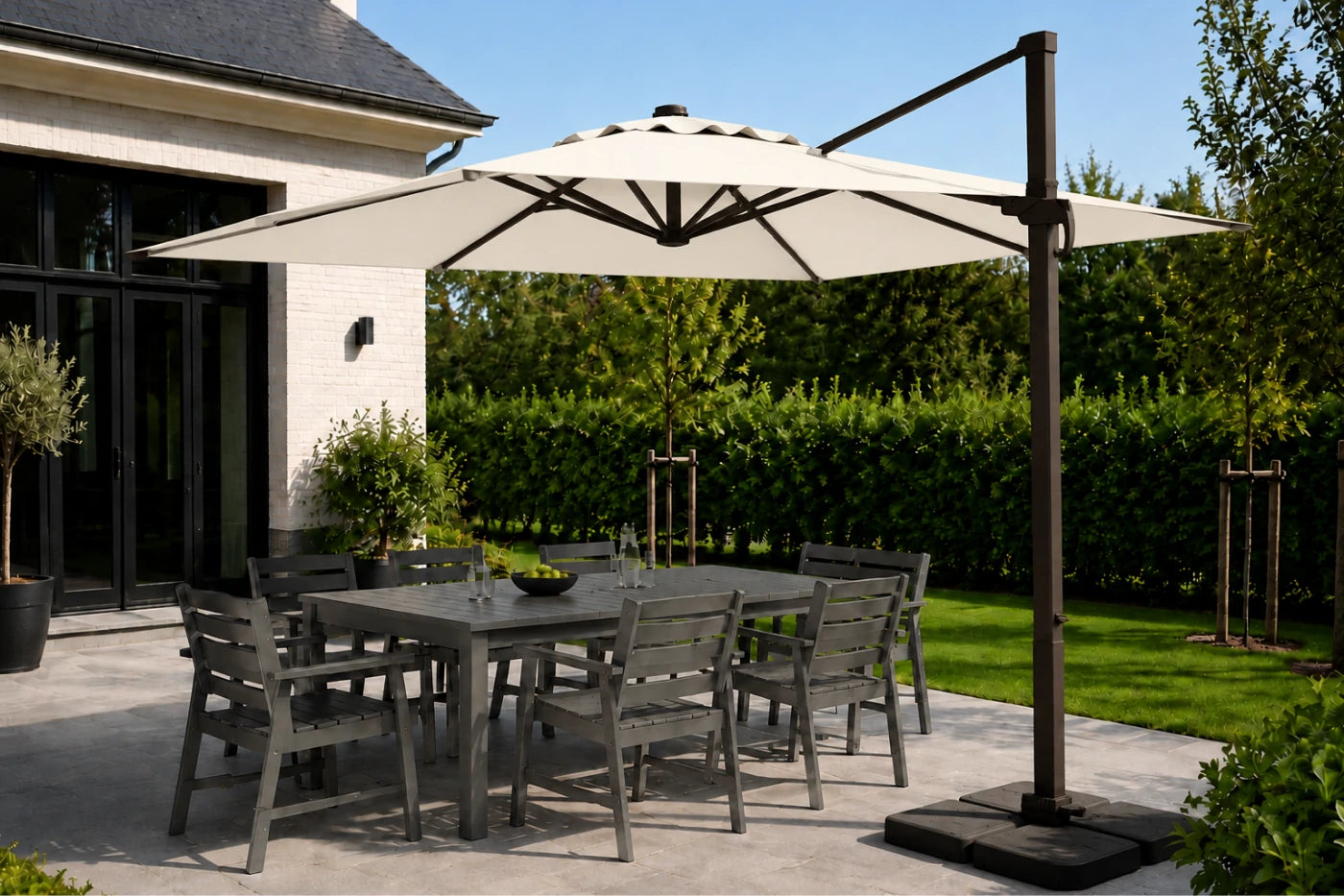 A large beige patio umbrella with a dark metal cantilever pole and base, shading a gray outdoor dining table and chairs on a patio. The background shows a modern house, green hedges, and a lawn under a clear blue sky.