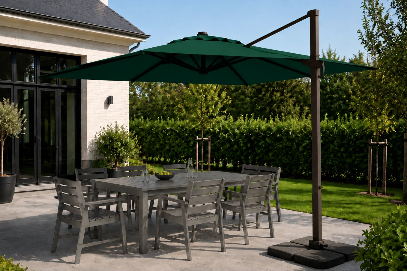 A dark green cantilever patio umbrella with a square base and brown metal pole, shading a gray outdoor dining table and chairs on a patio next to a house.