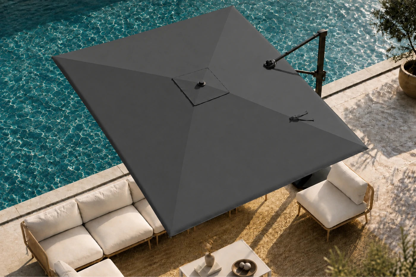 A large cantilever patio umbrella in dark gray shading an outdoor lounge area next to a swimming pool. The umbrella features a black metal support arm and is shown from an elevated angle over a beige stone patio with cream-colored outdoor furniture.