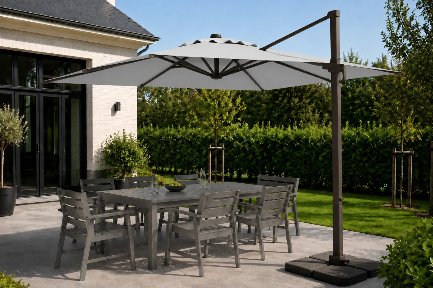 A large cantilever patio umbrella with a dark metal pole and base, shading a gray outdoor dining table and chairs on a paved patio. The scene is set in a landscaped backyard with green hedges, grass, and a house in the background.