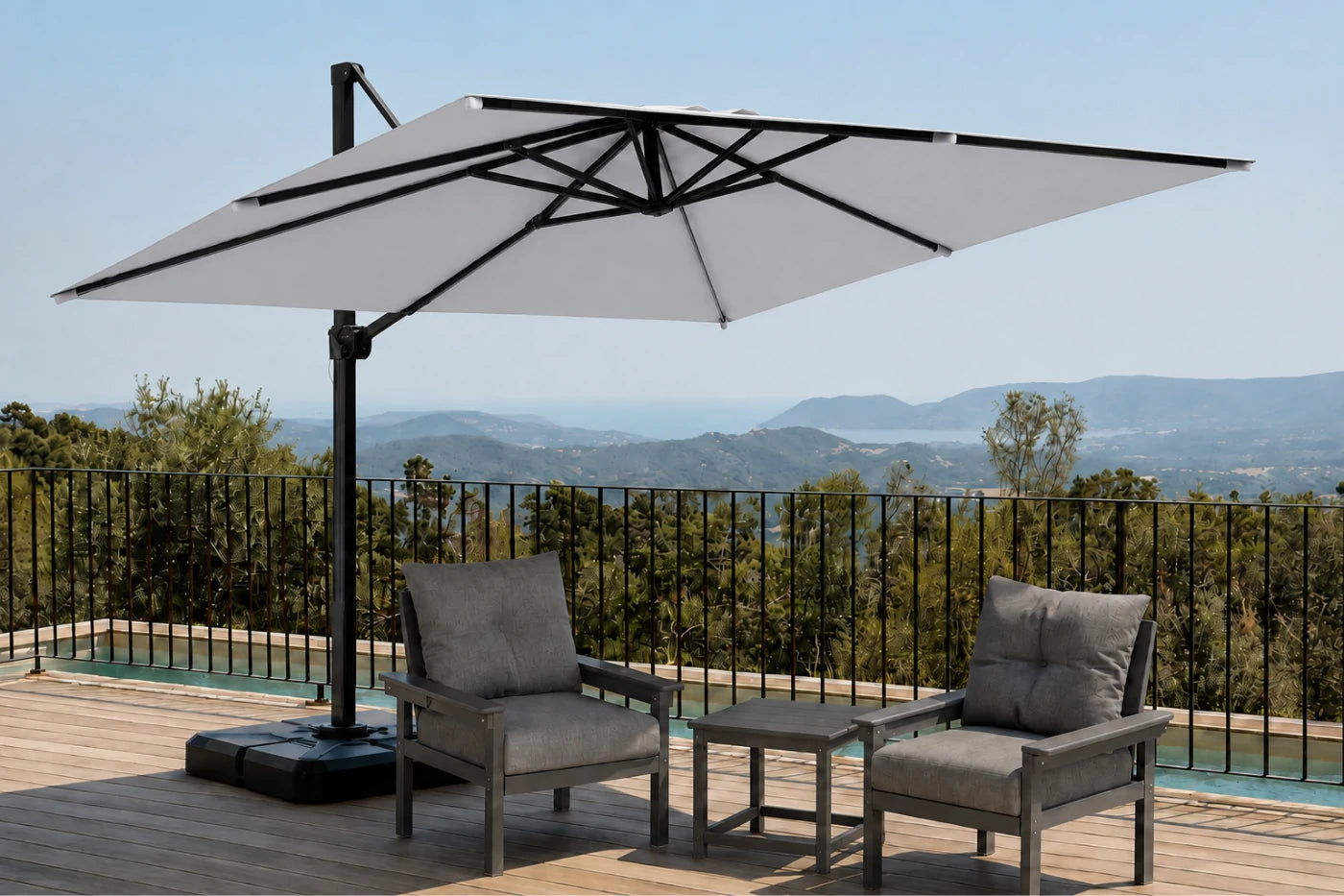 A large cantilever patio umbrella with a gray canopy and black frame is shown on a wooden deck beside a pool, shading two gray outdoor armchairs and a small side table. A real outdoor setting with a railing and distant hills is visible, so this is Lifestyle rather than Product/Packaging.