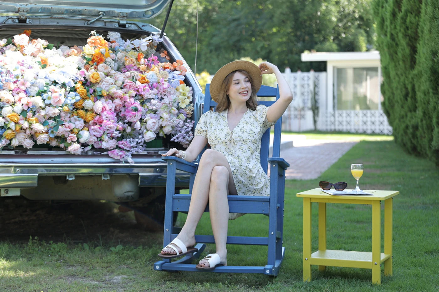 Woman sitting in a blue chair next to a truck filled with flowers, with a yellow table and sunglasses in the background.