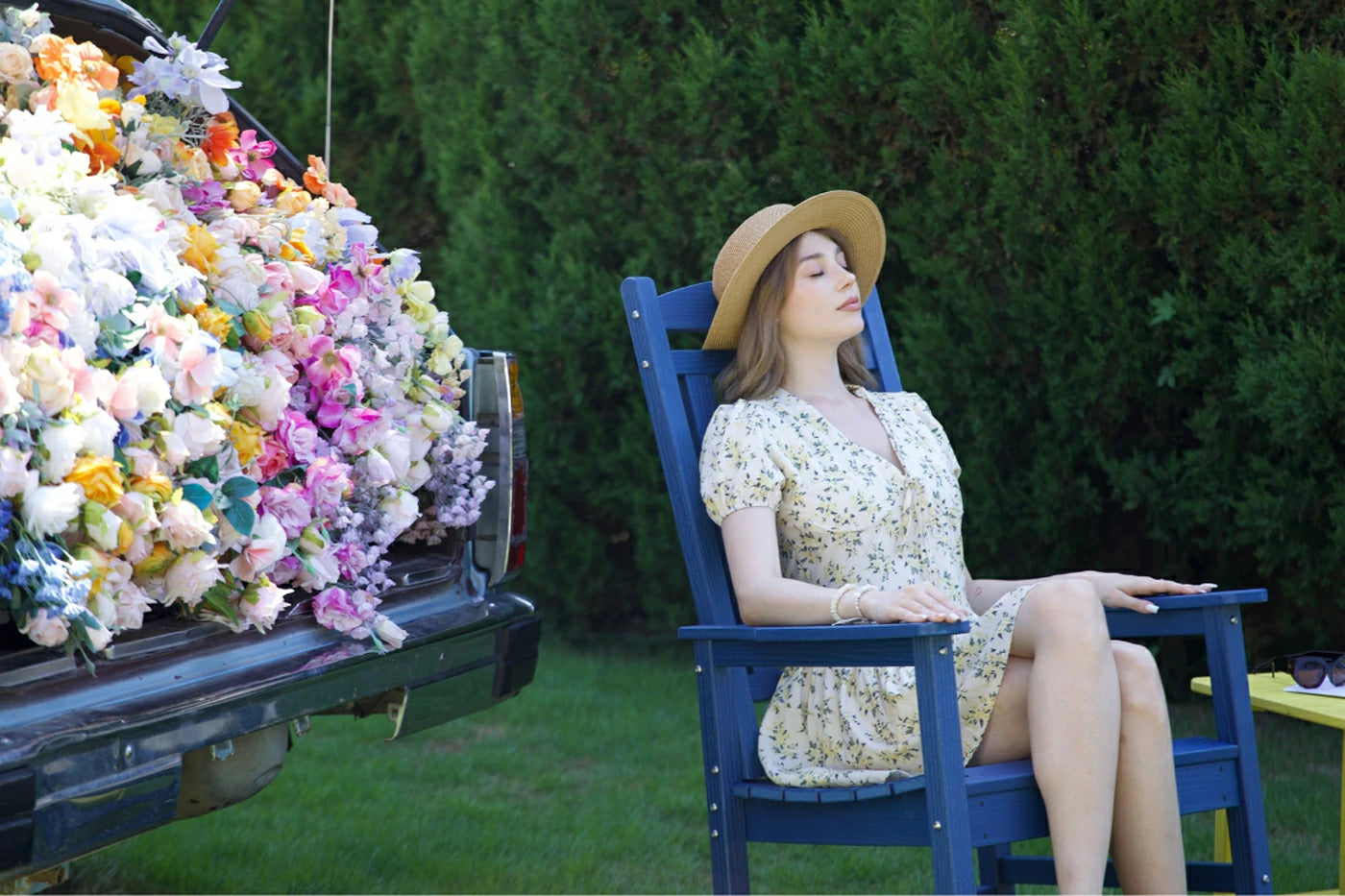 Woman in a floral dress sitting on a blue chair next to a truck filled with flowers.