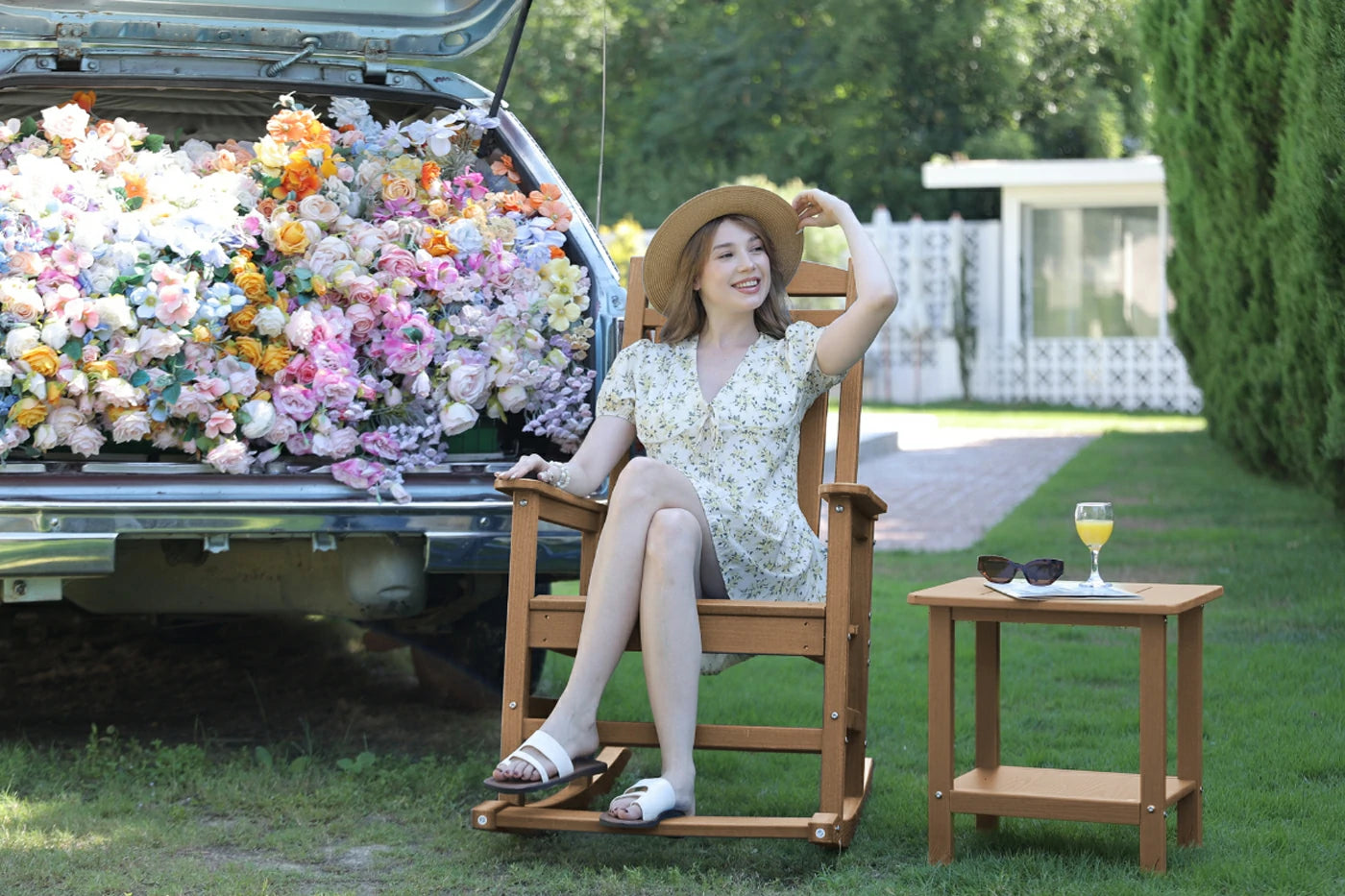 Woman sitting on a wooden chair with flowers in the back of a truck, enjoying a drink outdoors.