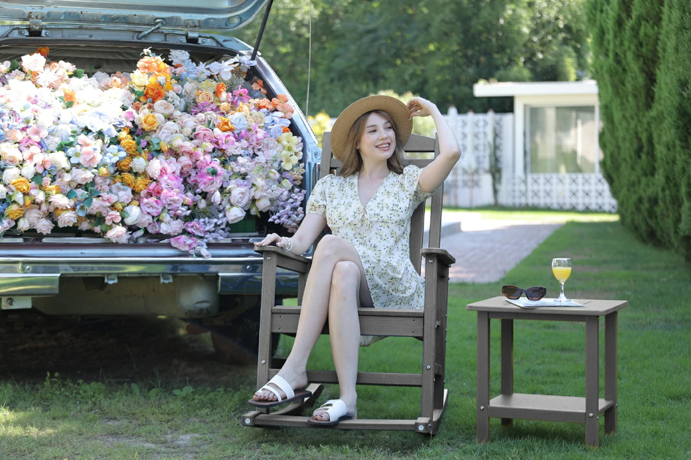 Woman sitting in a rocking chair next to a car filled with flowers, with a table and sunglasses in the foreground.