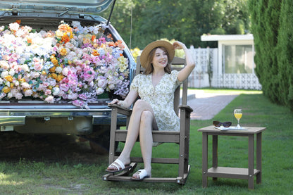 Woman sitting in a rocking chair next to a car filled with flowers, with a table and sunglasses in the foreground.
