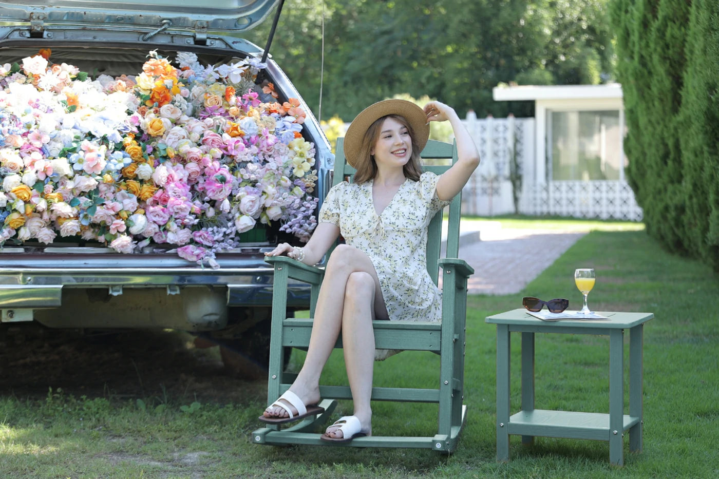 Woman sitting in a chair next to a car filled with flowers, with a table and sunglasses in the foreground.